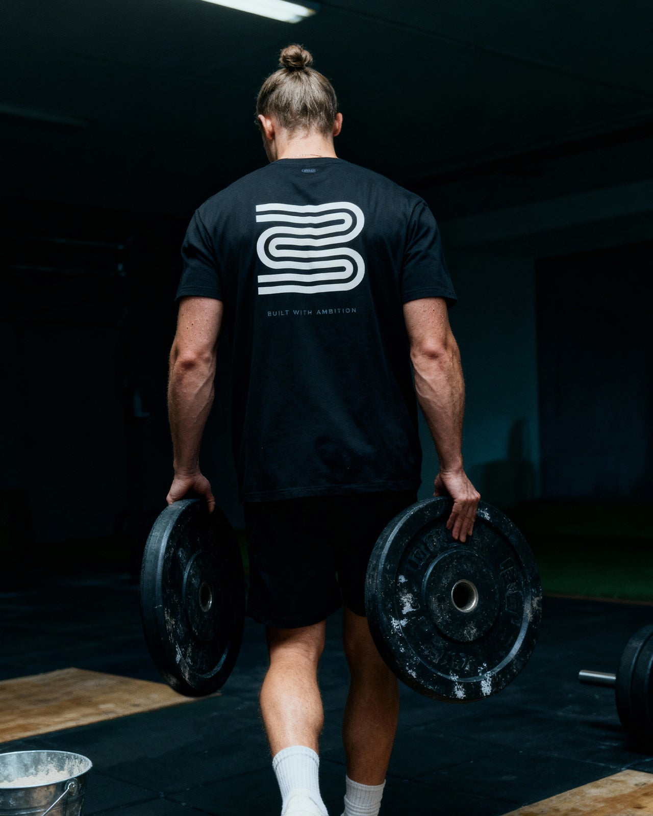 Athlete in gym carrying weight plates, wearing black training tee with ambition slogan